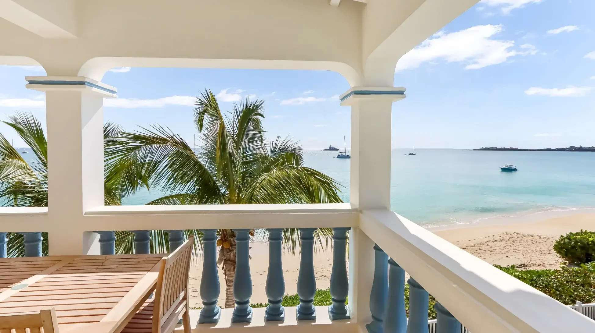 Your private oceanfront balcony features a wooden dining set beneath swaying palms, with white columns and blue balusters framing endless turquoise waters where sailboats drift by.