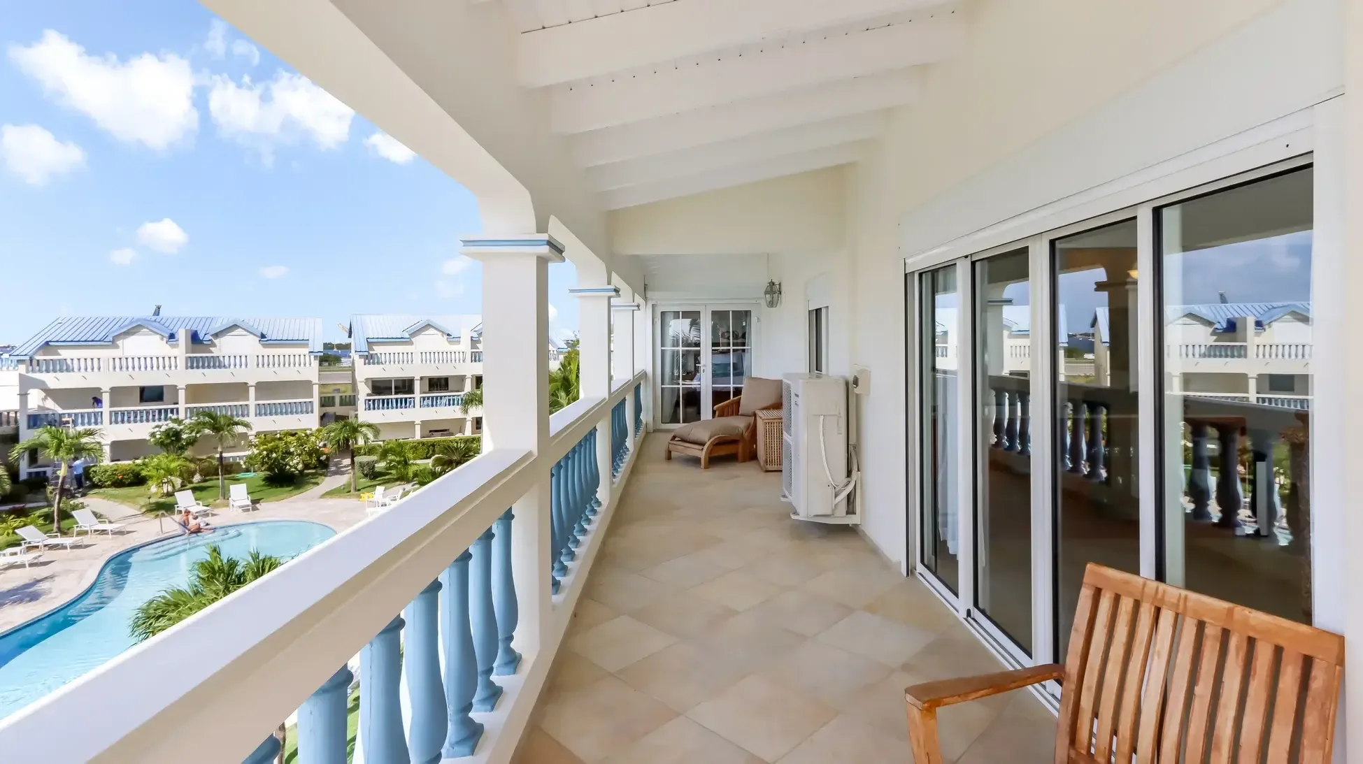 Covered balcony with tiled floor and lounge chair. View of the resort pool, palm trees, and neighboring buildings with blue roofs.