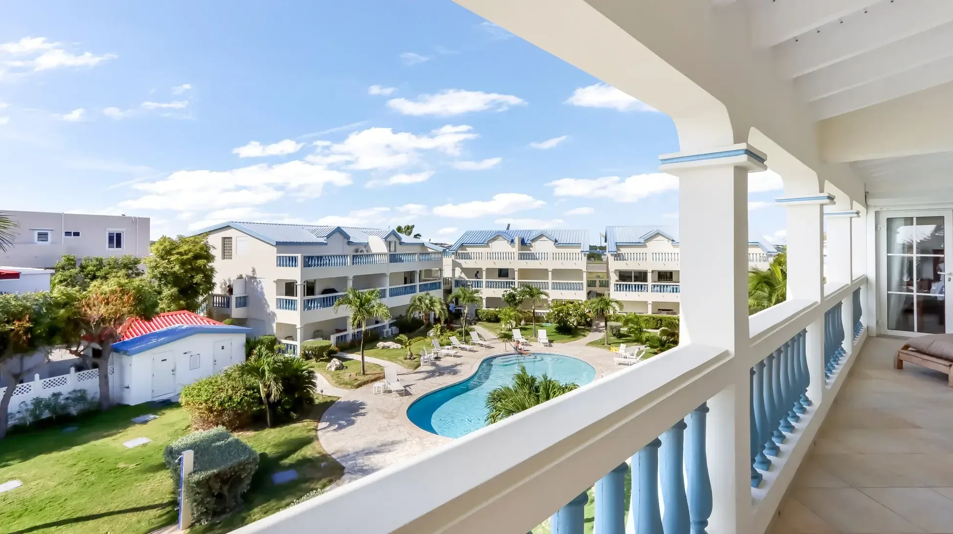 Blue balustrade frames the kidney-shaped pool with lounge chairs, manicured lawns, and white buildings with blue roofs under clear Caribbean skies.