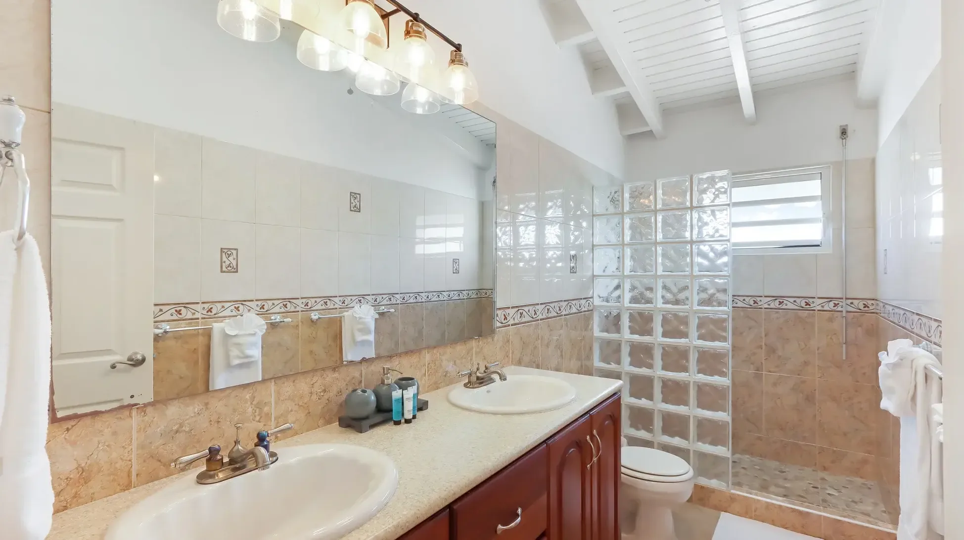 Double vanity with white sinks and wood cabinets. Glass block partition separates the shower area with decorative tile border.