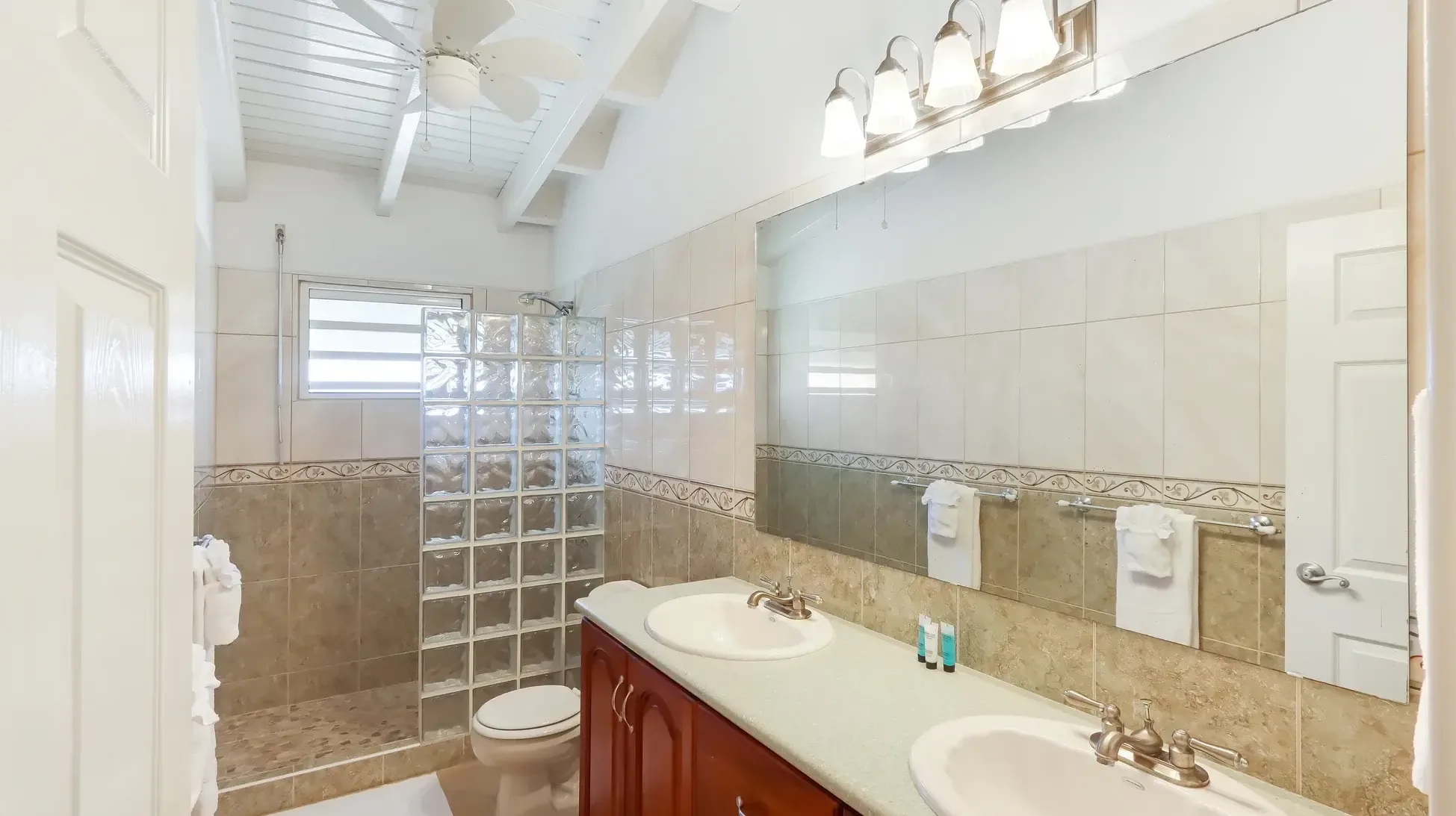 A modern bathroom with double vanity, walk-in shower featuring glass block wall and pebble floor. Vaulted ceilings add spaciousness.
