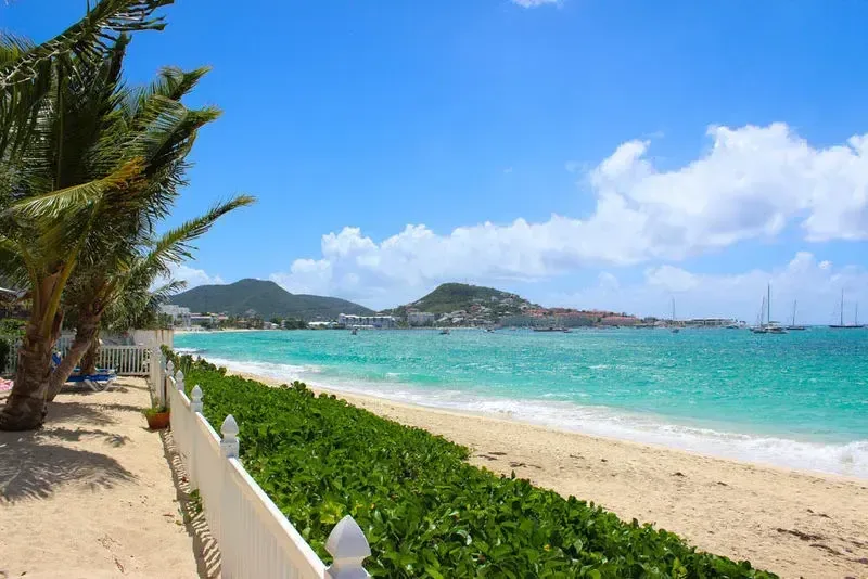 White sandy beach with turquoise ocean, palm trees shading blue lounge chairs. Sailboats drift in the distance with lush green hills.