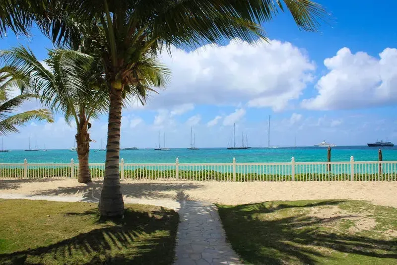 A stone walkway through grass leads to the beach. Palm trees frame the view of turquoise water with sailboats and a ship on the horizon.