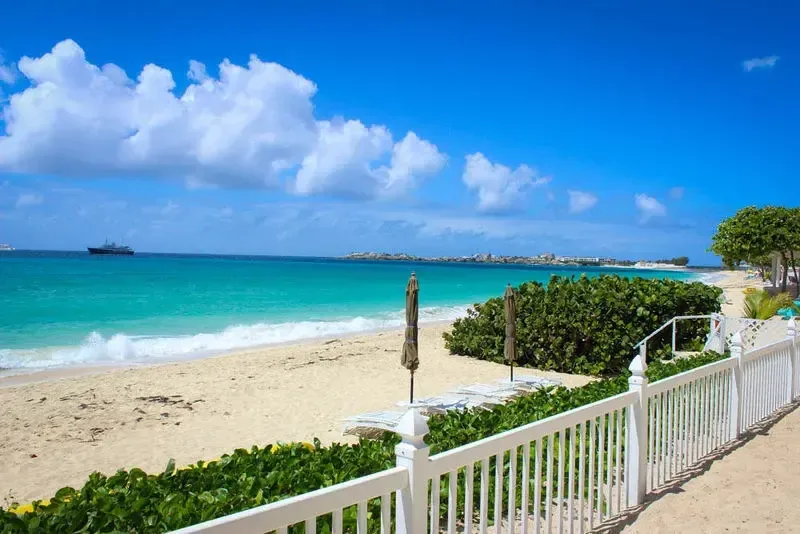 Turquoise ocean meets white sandy beach with lounge chairs and umbrellas. A white picket fence and tropical plants frame a large ship on the horizon.
