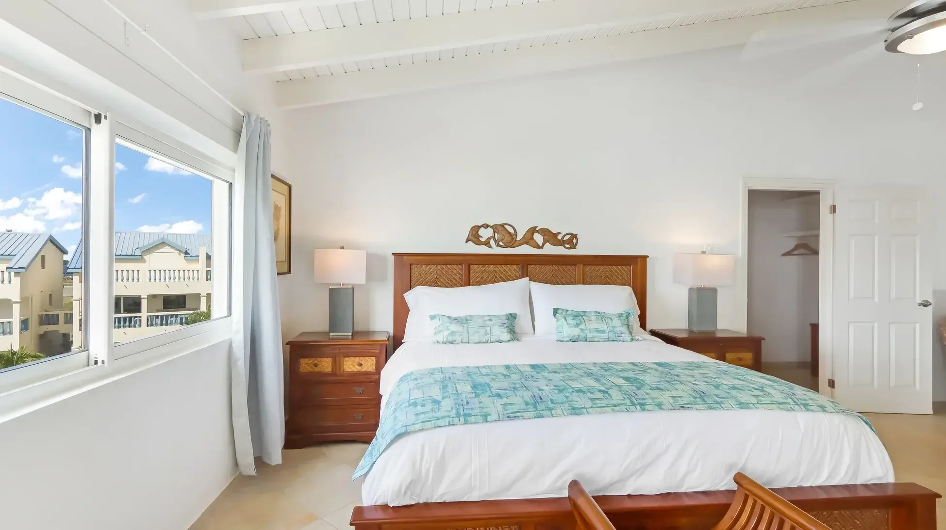 A king bed with woven headboard and dolphin carving. Modern lamps and vaulted ceiling frame windows with coastal views.