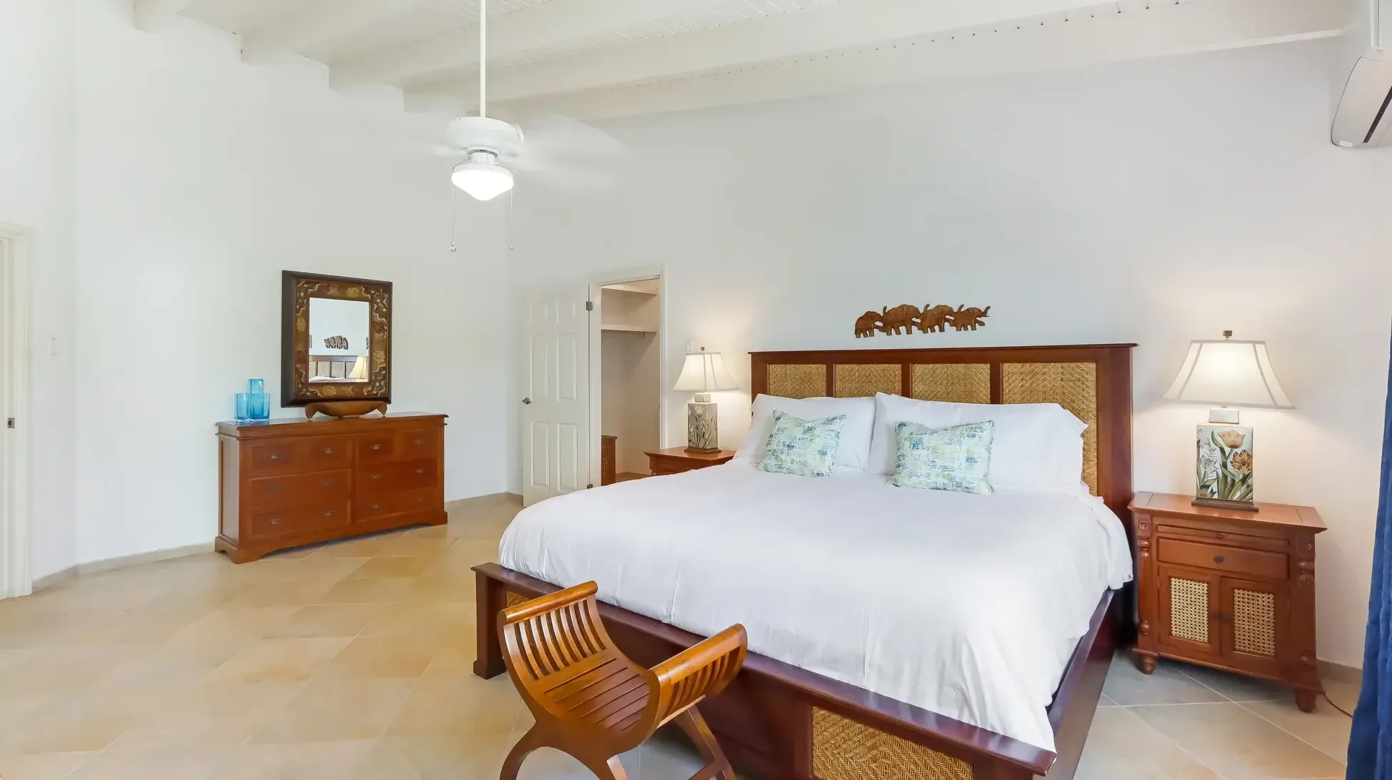 A large bed with dark wood and woven headboard, two bedside tables, wooden bench, and dresser with mirror. Blue glass accents add Caribbean charm.