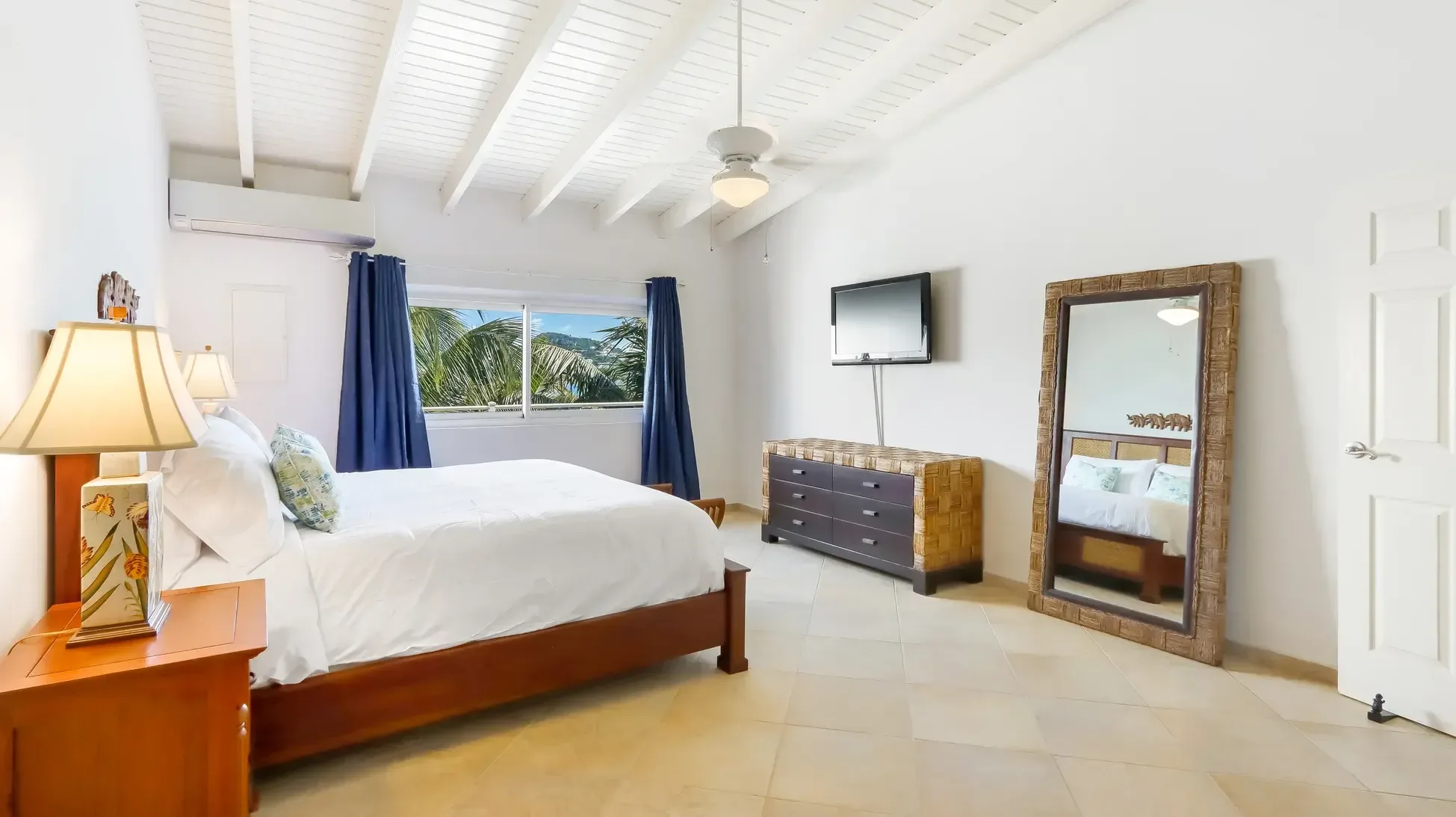 A bed with white linens and floral pillows, wall-mounted TV, and floor mirror. Vaulted ceiling and window with palm trees view.
