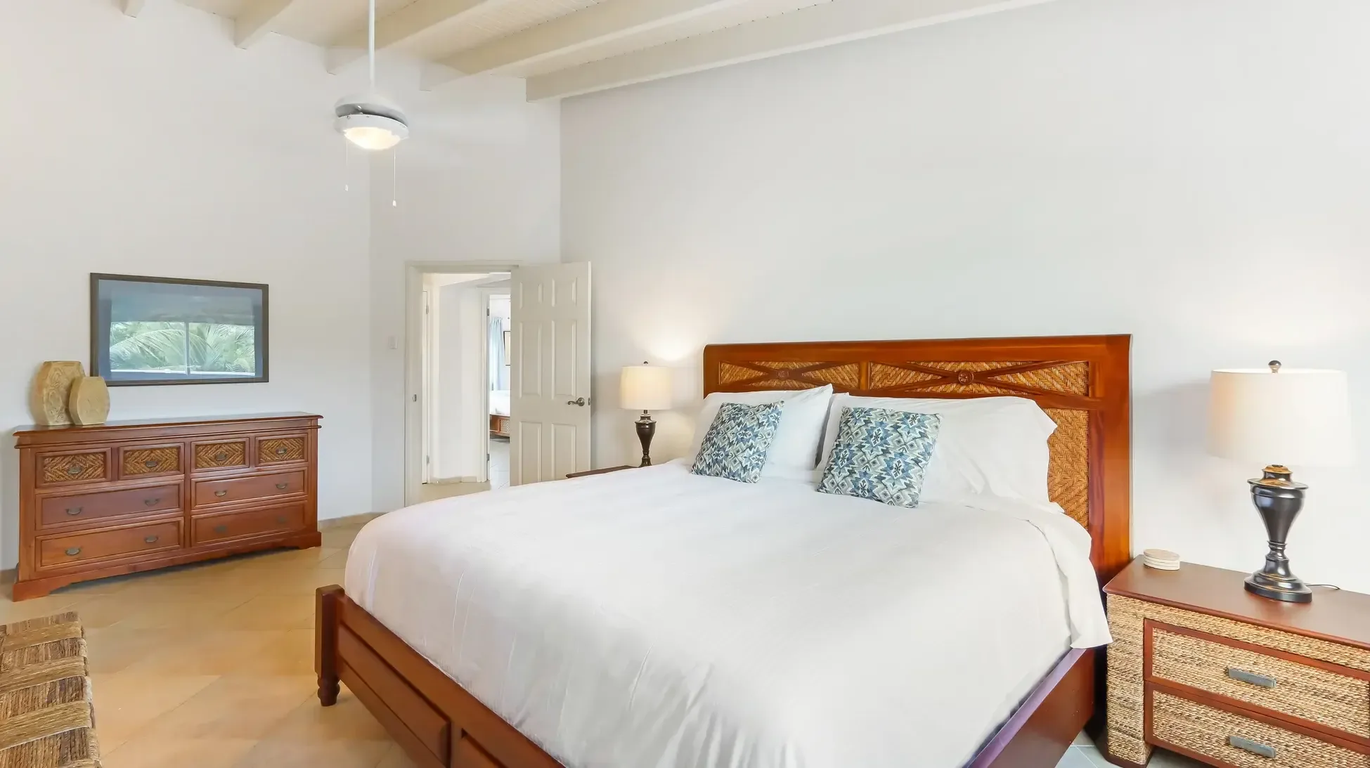 A king bed with wooden frame and woven headboard sits beneath slanted beamed ceilings. Two nightstands and a large dresser complete the space.