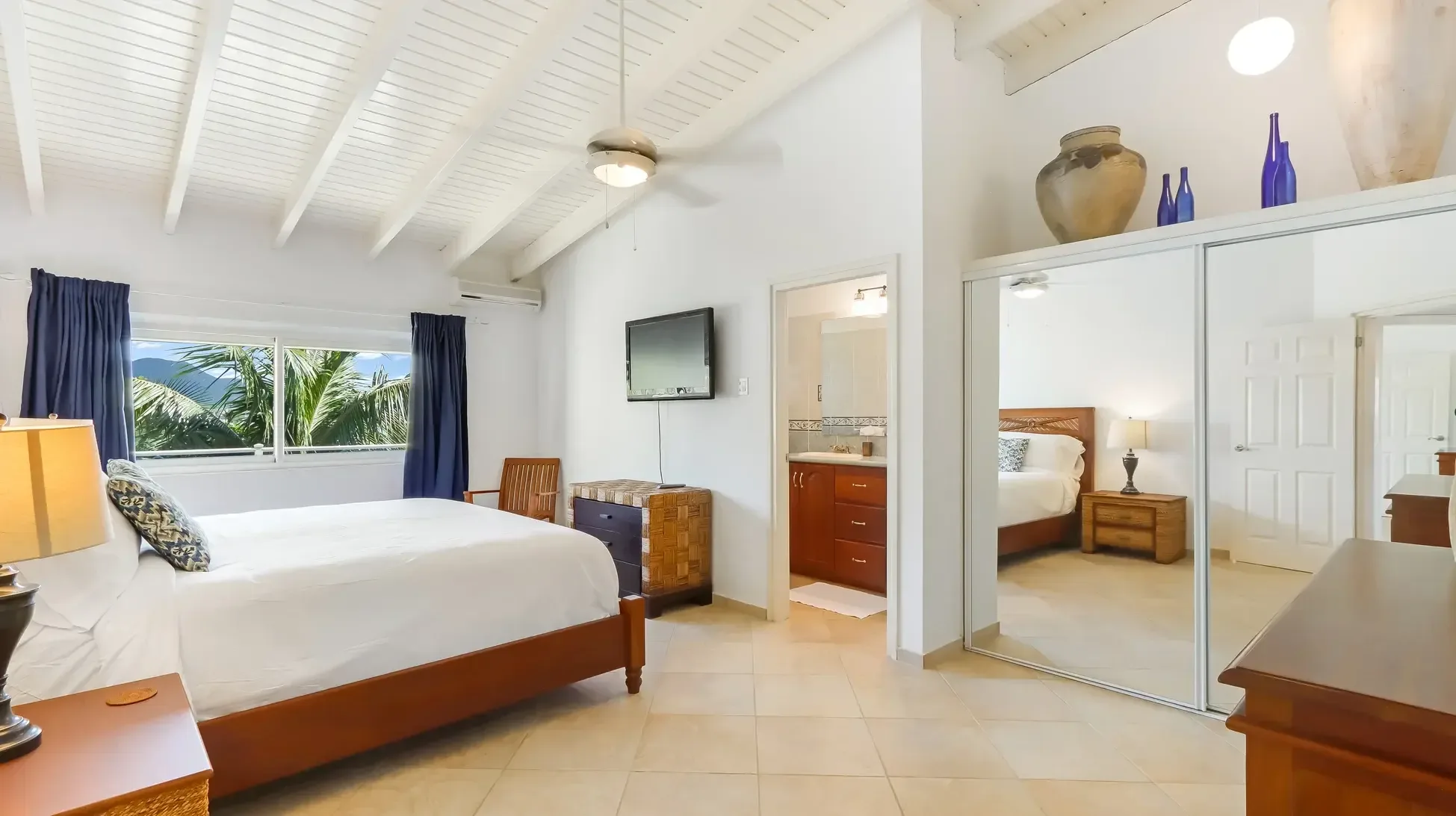 A spacious bedroom with wooden bed frame, TV, and ceiling fan. Blue curtains frame windows with tropical mountain views, and an ensuite bathroom is visible.