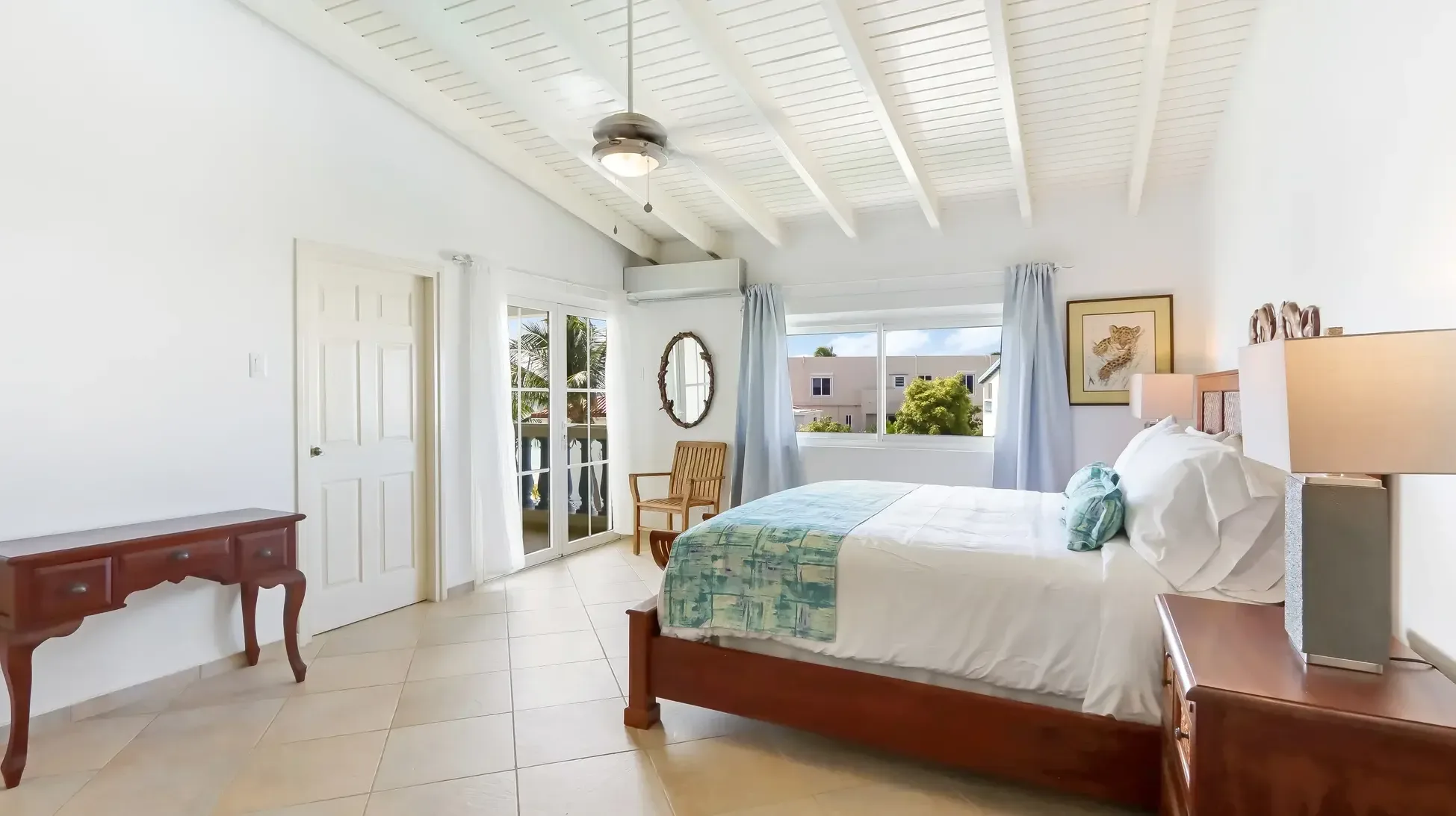 A cozy bedroom with wooden bed, white linens, and blue runner. Glass doors open to a private balcony with palm trees view.