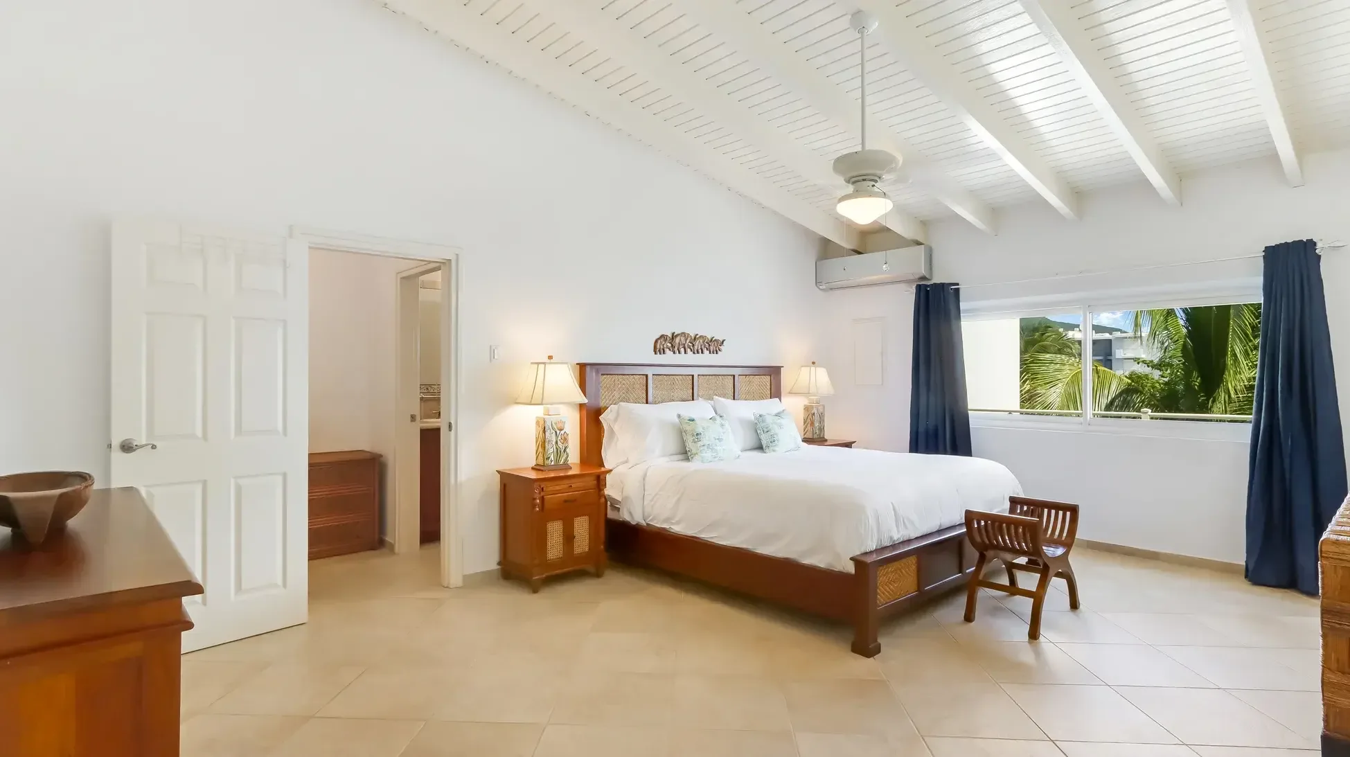 The primary bedroom features a wooden bed frame with woven headboard, white linens, and navy curtains. Vaulted ceilings and tropical views complete the retreat.
