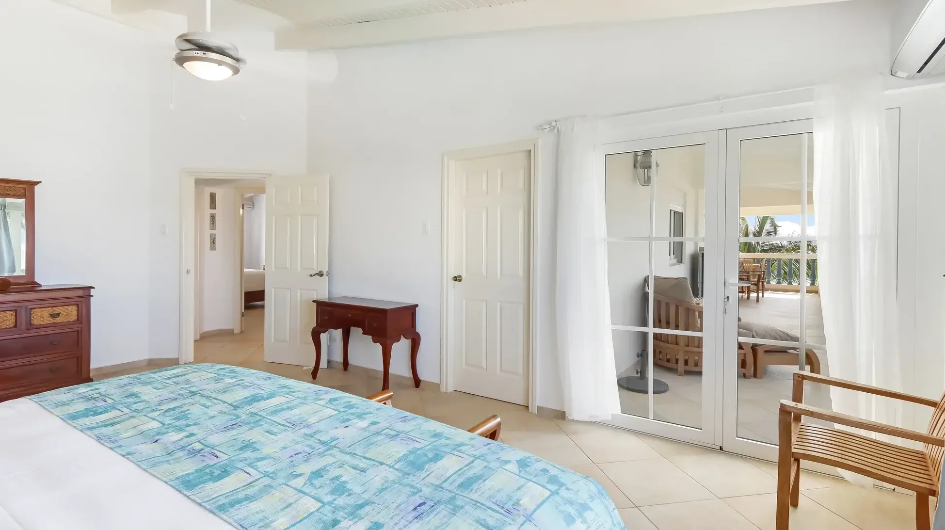 A king bed with blue comforter and dark wood dresser. Sliding doors lead to a furnished terrace with ocean views.