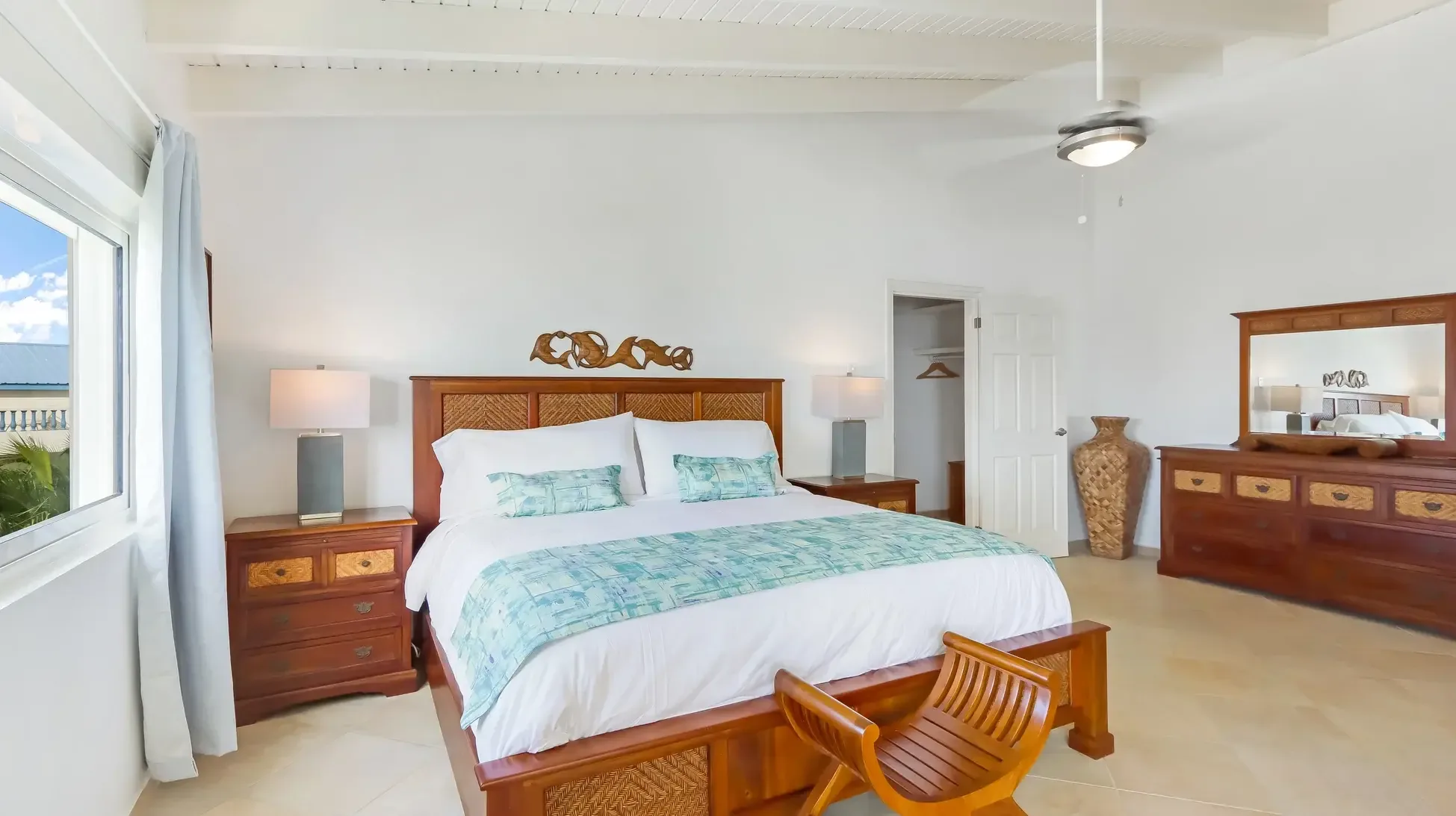 A king bed with tropical bedding and carved wooden headboard. Nightstands with lamps and a curved wooden bench add elegance.