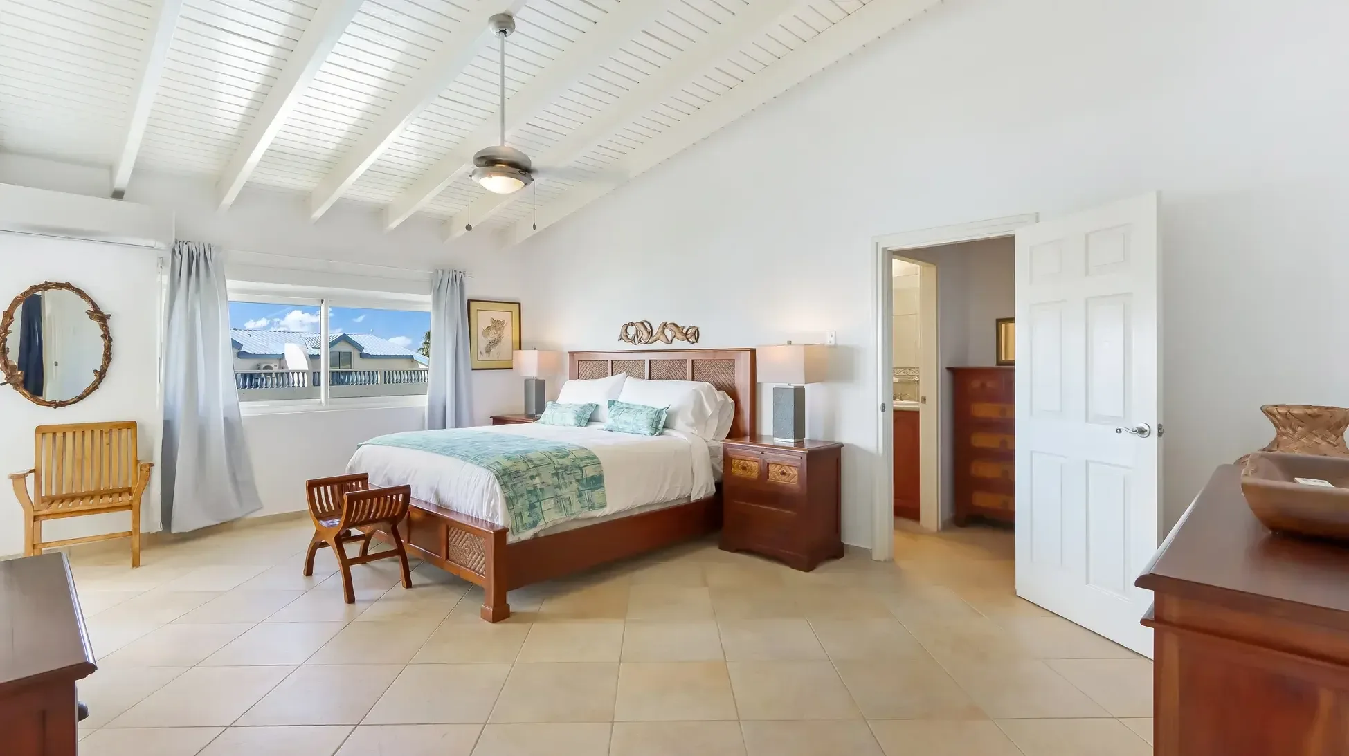 A serene bedroom with wooden bed, decorative headboard, armchair, and vaulted beamed ceiling. Grey curtains frame the windows.