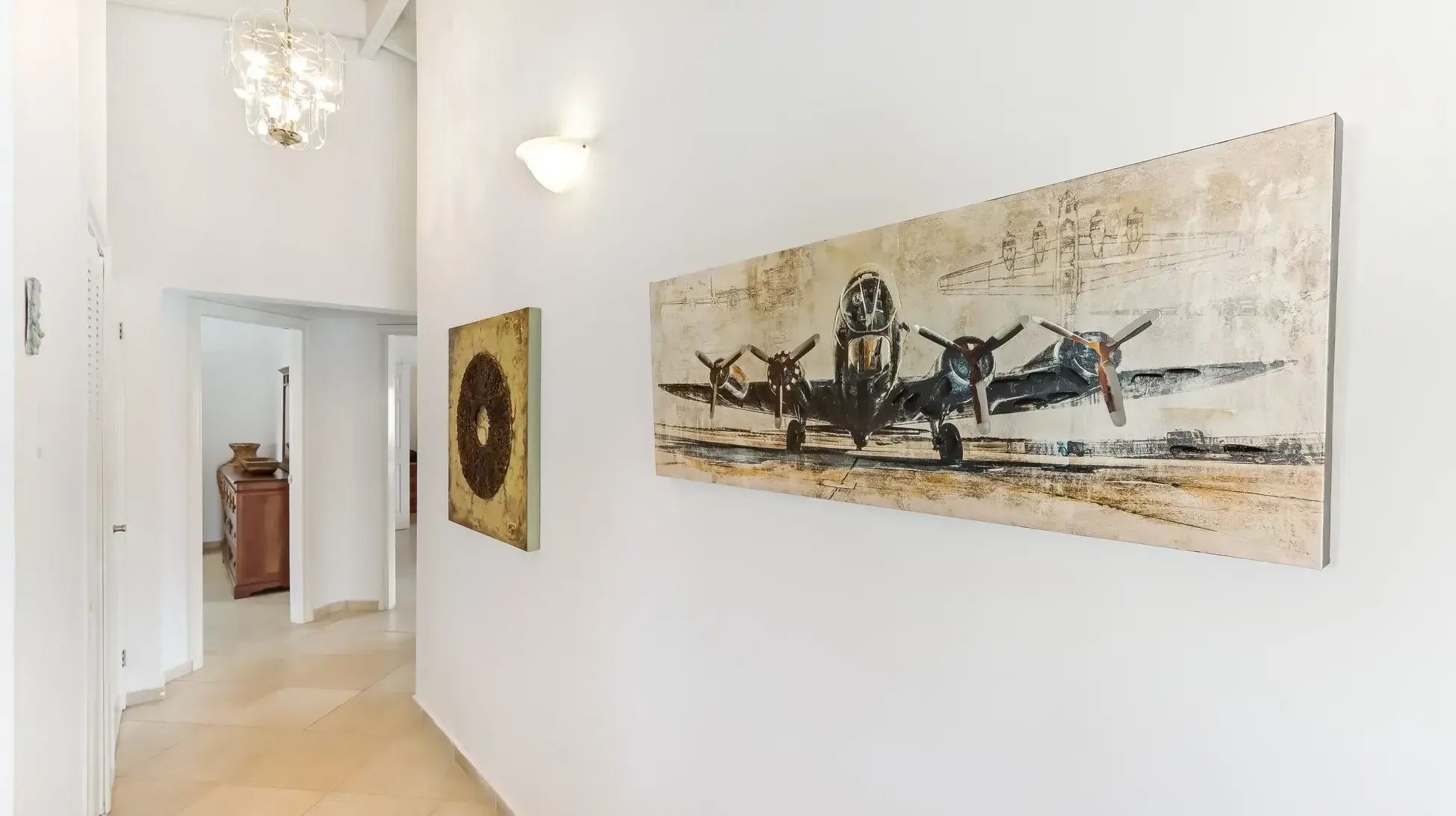 A gallery hallway featuring vintage airplane painting and abstract art, with wall sconces and chandelier illuminating the tile flooring.