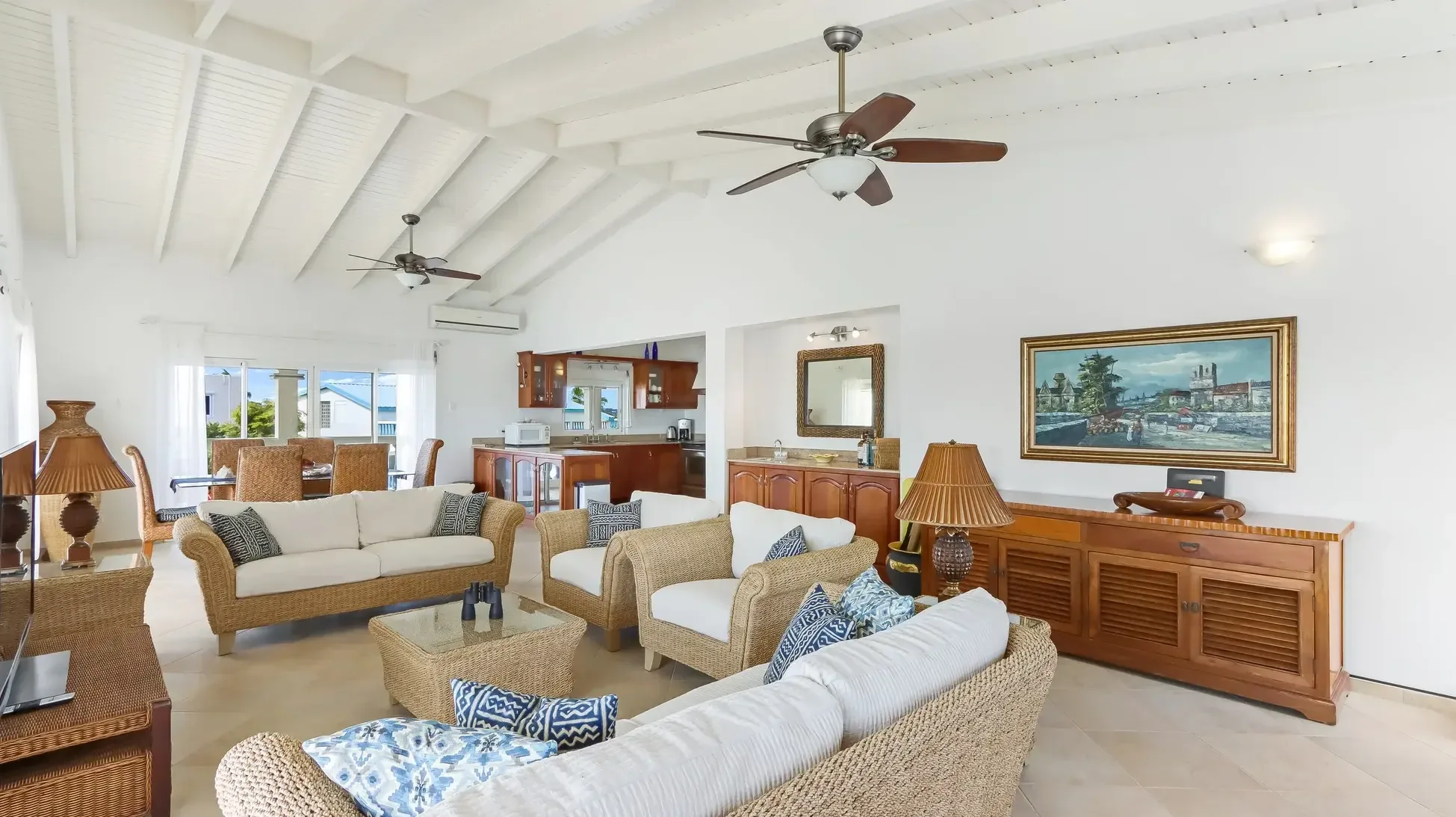 The open living and dining area showcases wicker armchairs, a glass coffee table, and a large wooden sideboard beneath vaulted ceilings with tropical views.