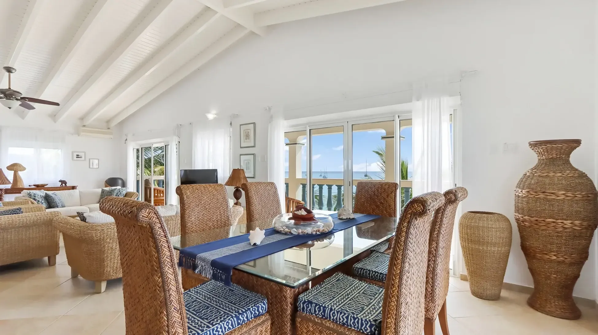Open-plan living with nautical-themed decor, wicker furniture, and vaulted ceilings. Large sliding doors showcase sailboats on the horizon.