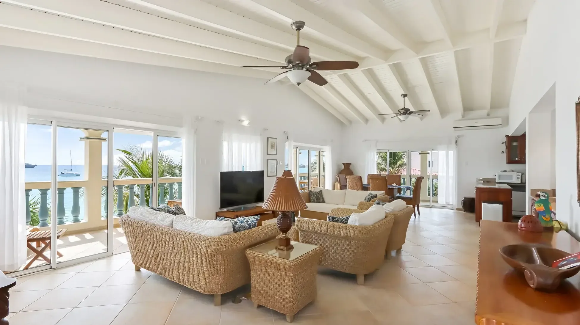 An open-concept living room where wicker furniture and vaulted ceilings meet panoramic ocean views. A catamaran glides past your sliding glass doors.