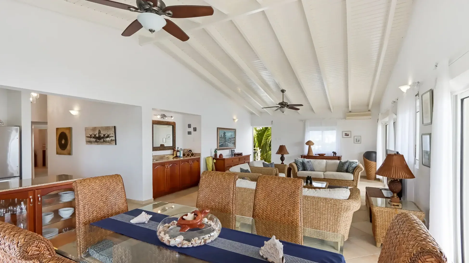 Open living space with dining area, wicker furniture, and built-in bar with glassware. Tropical foliage frames the sliding doors.