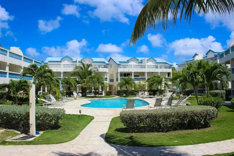 Kidney-shaped pool with paved sun deck and white lounge chairs. Multi-story white buildings with blue roofs and manicured hedges complete the scene.