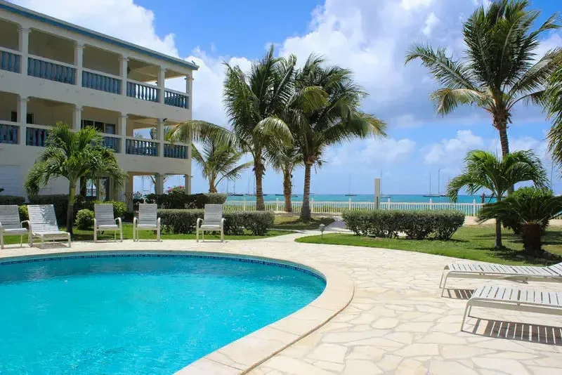 A blue curved pool with stone deck and white lounge chairs. The three-story building with balconies overlooks palm trees and ocean with sailboats.