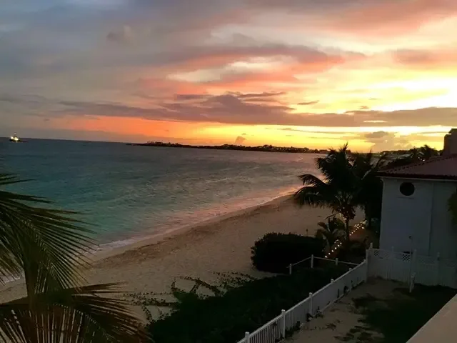 Orange, pink, and yellow clouds crown the turquoise ocean at sunset. Palm fronds and white picket fence frame a small boat on the horizon.
