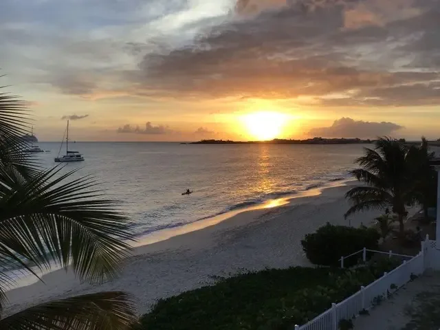 A dramatic sunset paints the sky as someone kayaks past white sandy shores. Palm trees and sailboats silhouette against the orange horizon.