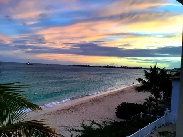 Sandy beach at sunset with gentle waves and pink-orange sky. Palm fronds sway as someone walks along the shoreline near the white picket fence.