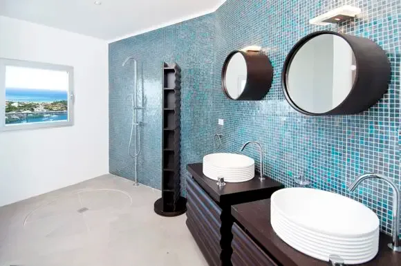 A spa-inspired double vanity features blue mosaic tile accents, round vessel sinks with matching mirrors, and a window framing the ocean vista.                                                                                                                                                                                                                                                                                                                                                                         +
 