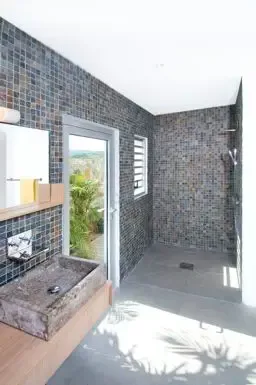 Natural stone vessel sink on wooden vanity contrasts with dark mosaic tile walls, while glass doors open to a lush outdoor shower area.                                                                                                                                                                                                                                                                                                                                                                                  +
 