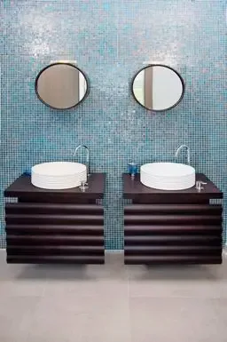 White vessel sinks rest on a dark wood floating vanity, complemented by round mirrors and a striking blue mosaic tile feature wall.                                                                                                                                                                                                                                                                                                                                                                                     +
 