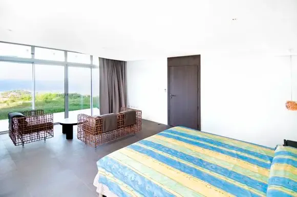 A spacious bedroom offers floor-to-ceiling ocean views, woven seating area, and a colorful striped bedspread creating a relaxed island atmosphere.                                                                                                                                                                                                                                                                                                                                                                             +
 
