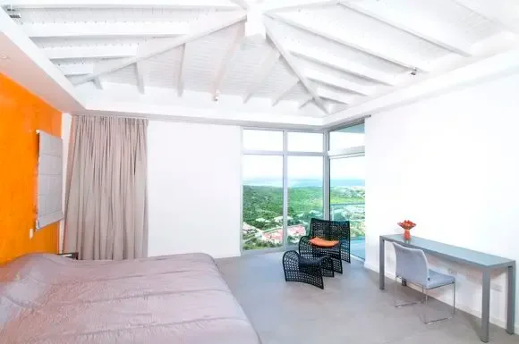 A vibrant orange accent wall anchors this bedroom with vaulted ceiling, floor-to-ceiling windows, woven lounge chair, and stunning landscape views.                                                                                                                                                                                                                                                                                                                                                                      +
 