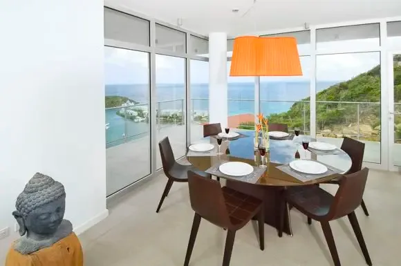 A round glass-top table set for six catches light from the oversized orange pendant, while floor-to-ceiling windows showcase the panoramic ocean view and Buddha statue accent.                                                                                                                                                                                                                                                                                                                                         +
 