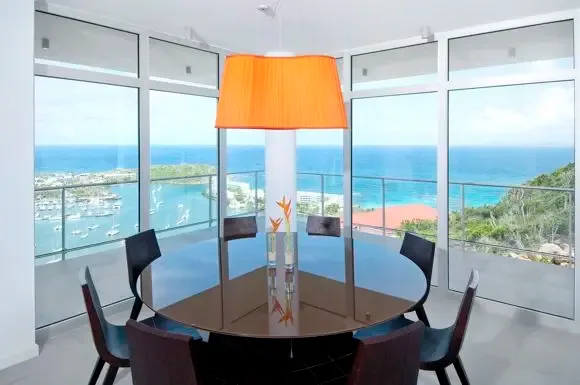 Modern dining elegance with round glass table, oversized orange pendant light, and expansive windows revealing the harbor and Caribbean Sea beyond.                                                                                                                                                                                                                                                                                                                                                                       +
 