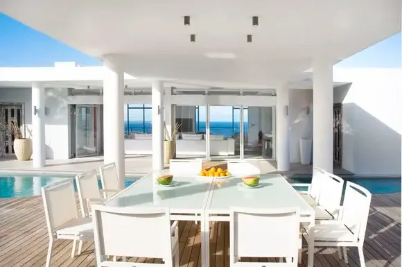 An elegant white outdoor dining set rests on warm wood decking, offering pool and ocean views through minimalist architecture and glass railings.                                                                                                                                                                                                                                                                                                                                                                        +
 