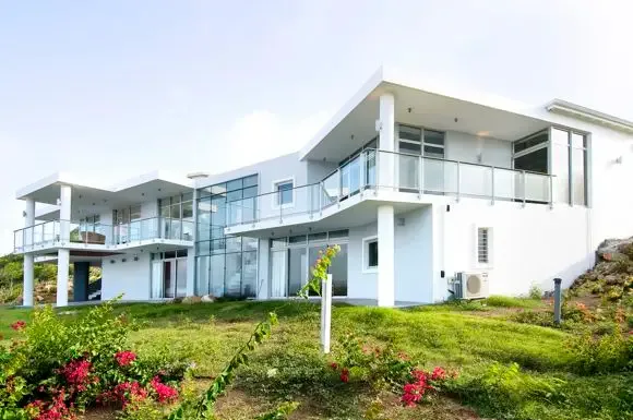 A striking aerial perspective showcases the villa's modern multi-level architecture, glass railings, and floor-to-ceiling windows framed by vibrant pink bougainvillea and lush hillsides.                                                                                                                                                                                                                                                                                                                       +
 