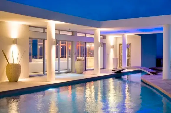 The villa's clean white lines and circular columns frame the illuminated infinity pool and decorative bridge, accented by warm wall sconces and cool blue recessed lighting at dusk.                                                                                                                                                                                                                                                                                                                                    +
 