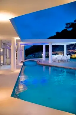 The illuminated swimming pool and covered terrace create a stunning nighttime tableau, with white columns and warm lighting under the twilight sky.                                                                                                                                                                                                                                                                                                                                                                      +
 