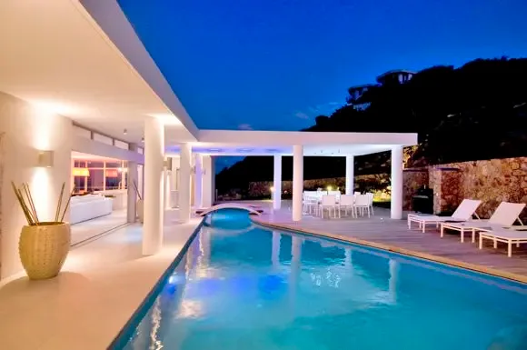 A magical twilight scene captures the long illuminated pool, covered outdoor dining colonnade, and sun loungers under a deep blue Caribbean sky.                                                                                                                                                                                                                                                                                                                                                                      +
 