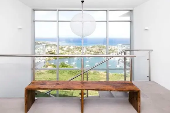 A minimalist interior corridor features a rustic wooden bench beneath a dramatic sphere pendant, with floor-to-ceiling windows showcasing the coastal harbor panorama.                                                                                                                                                                                                                                                                                                                                                     +
 