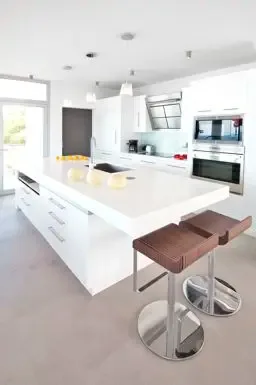 The sleek kitchen island with built-in sink and wooden bar stools anchors this minimalist white space, equipped with high-end stainless steel appliances.                                                                                                                                                                                                                                                                                                                                                                +
 