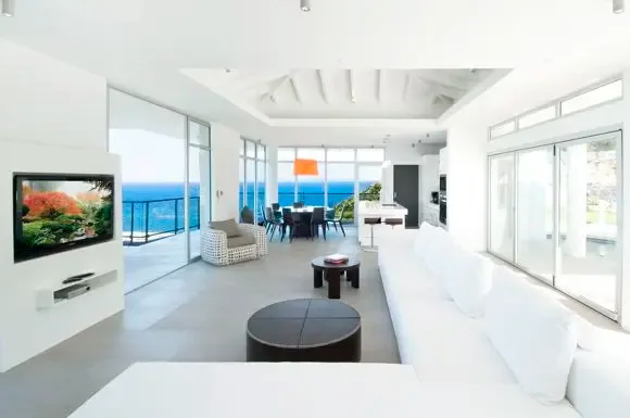A bright open-concept living area features a sprawling white sectional, vaulted white-beam ceilings, and floor-to-ceiling windows framing panoramic ocean views.                                                                                                                                                                                                                                                                                                                                                            +
 