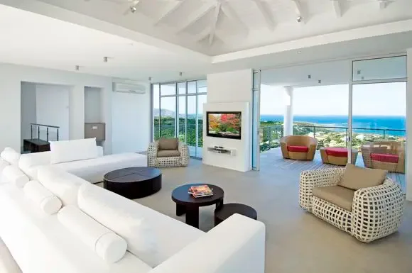 Modern living space with white sectional, circular coffee tables, and woven armchair flows through sliding glass doors to a terrace with red-cushioned seating and ocean views.                                                                                                                                                                                                                                                                                                                                          +
 
