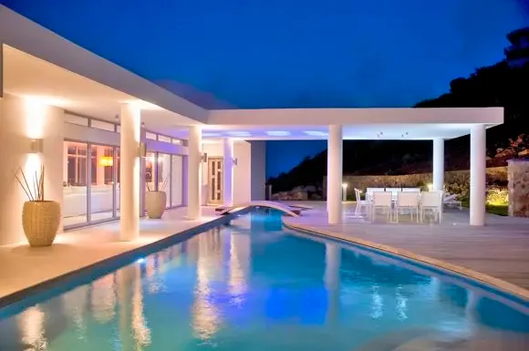 Evening elegance defines this scene: a long lit swimming pool with small bridge, white-columned terrace with outdoor dining, and the last light of dusk.                                                                                                                                                                                                                                                                                                                                                                      +
 
