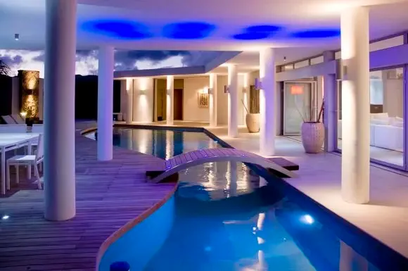Night transforms the villa as the pool flows through the terrace, illuminated by blue ceiling lights and elegant white columns against the twilight sky.                                                                                                                                                                                                                                                                                                                                                                   +
 