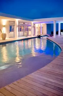A curved swimming pool glows against the wooden deck and modern white architecture, while interior lights create a warm invitation through glass doors.                                                                                                                                                                                                                                                                                                                                                                        +
 