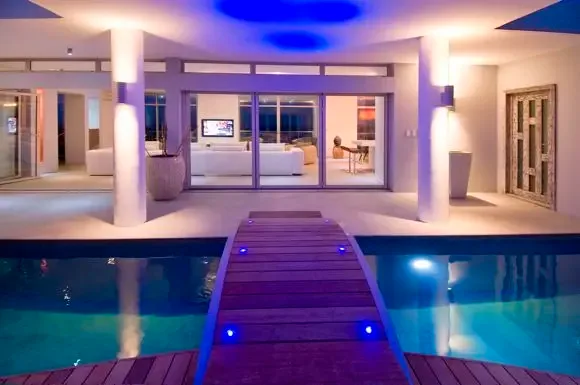 Blue LED lights illuminate the wooden bridge and pool at night, while dramatic ceiling lighting showcases the open-plan living room visible through glass doors.                                                                                                                                                                                                                                                                                                                                                             +
 