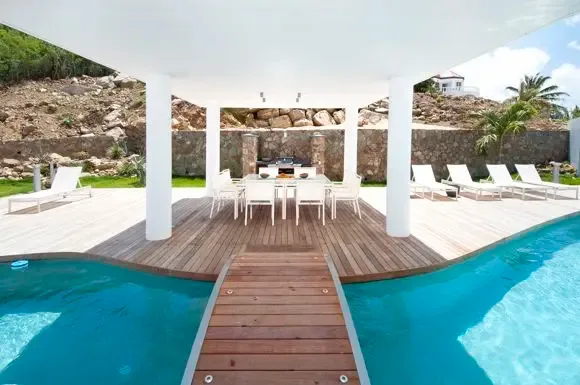 A covered outdoor kitchen and dining area sit beside the pool, accessible via a wooden bridge, with modern white architecture contrasting natural stone walls.                                                                                                                                                                                                                                                                                                                                                          +
 