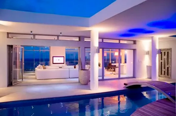 The infinity-style pool glows blue at dusk, its curved wooden bridge leading to the modern living area where white furniture reflects ambient lighting.                                                                                                                                                                                                                                                                                                                                                                   +
 